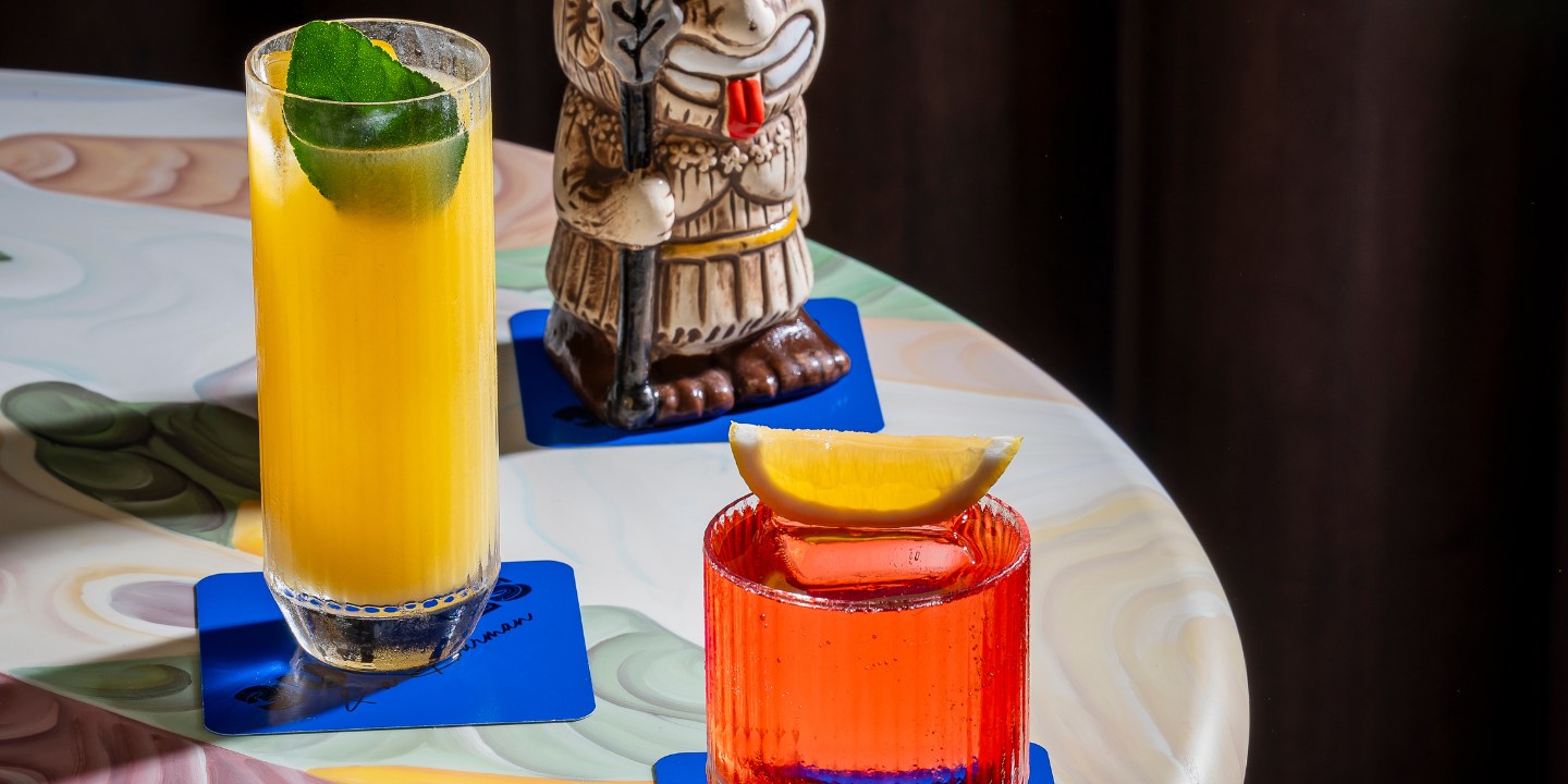 Three colorful cocktails on a swirled pattern table. Left: yellow drink with lime in a tall glass, middle: ornate tiki mug, right: red drink with lemon.